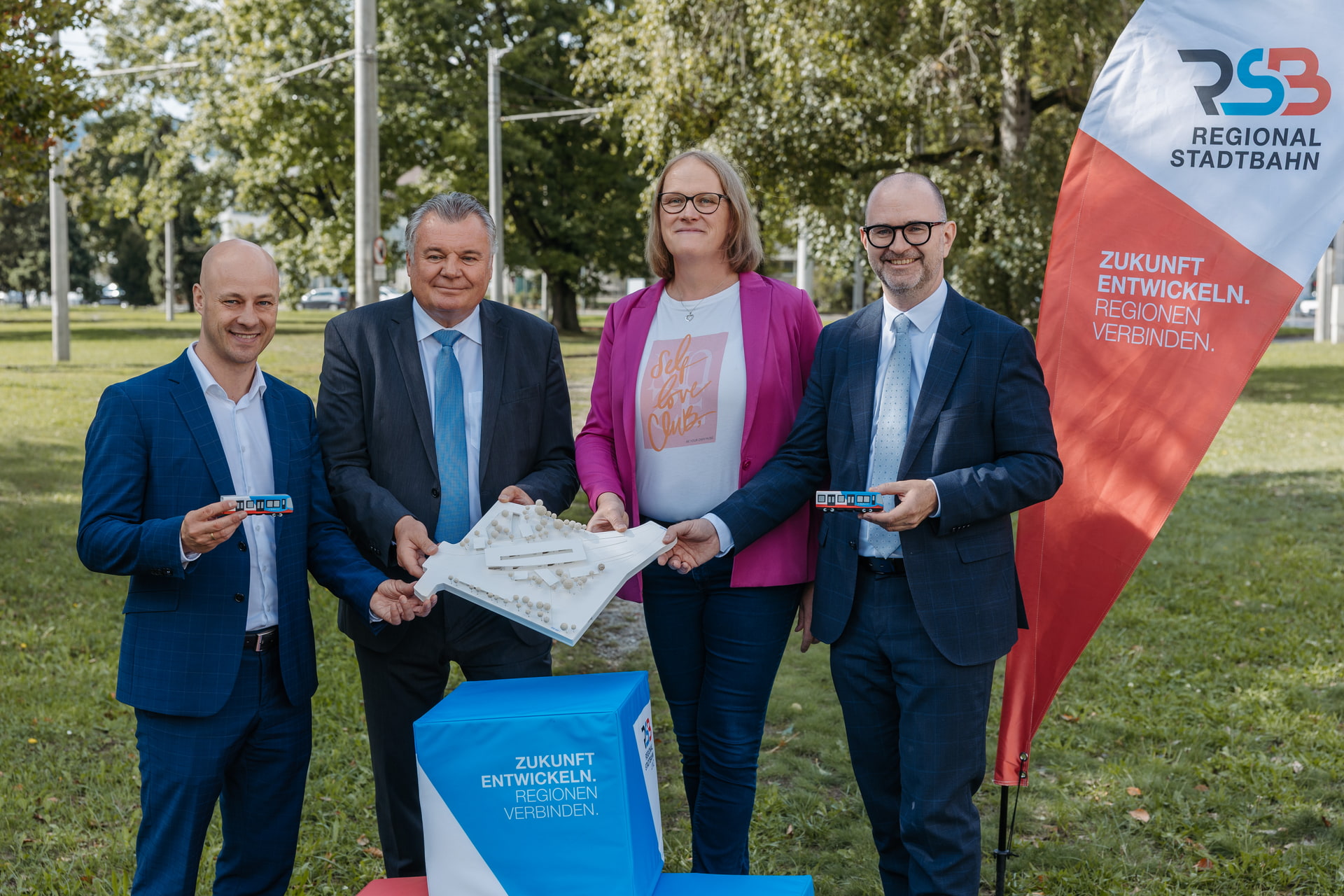 Eröffnung RSB Infopark Donaubahnhof Martin Hajart (Vizebürgermeister der Stadt Linz), Günther Steinkellner (Landesrat für Mobilität und Infrastruktur), Sonja Wiesholzer (Geschäftsführerin der Schiene OÖ) und Dietmar Prammer (Bürgermeister der Stadt Linz) halten Bahnmodell; Banner mit Regionalbahn und Motto Zukunft entwickeln, Regionen verbinden.